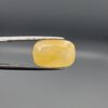 Studio photograph held in steel tweezers showcasing a large 13.70 carat natural cushion-shaped yellow sapphire (Pukhraj) loose gemstone with vibrant golden-yellow hue.