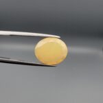 Studio photograph held in steel tweezers showcasing a 4.05 carat natural oval-shaped Ethiopian opal loose gemstone featuring a double-side fancy cut with flat and domed facets.