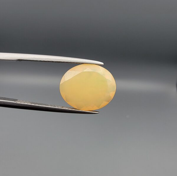 Studio photograph held in steel tweezers showcasing a 4.05 carat natural oval-shaped Ethiopian opal loose gemstone featuring a double-side fancy cut with flat and domed facets.