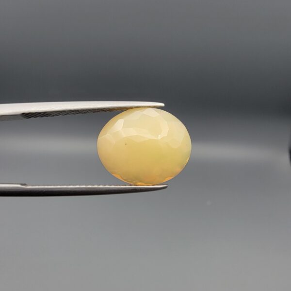 Studio photograph held in steel tweezers showcasing a 4.05 carat natural oval-shaped Ethiopian opal loose gemstone featuring a double-side fancy cut with flat and domed facets.
