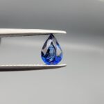 Studio photograph held in steel tweezers showcasing a 1.06 carat natural pear-shaped blue sapphire (Neelam) loose gemstone with exceptional luster and classic blue facets.