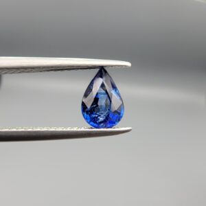 Studio photograph held in steel tweezers showcasing a 1.06 carat natural pear-shaped blue sapphire (Neelam) loose gemstone with exceptional luster and classic blue facets.