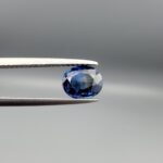 Studio photograph held in steel tweezers showcasing a 1.42 carat natural oval-shaped blue sapphire loose gemstone featuring classic blue facets and a natural inclusion pocket on the back.