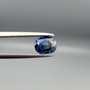 Studio photograph held in steel tweezers showcasing a 1.42 carat natural oval-shaped blue sapphire loose gemstone featuring classic blue facets and a natural inclusion pocket on the back.