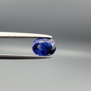 Studio photograph held in steel tweezers showcasing a 1.20 carat natural oval-shaped blue sapphire loose gemstone with rich blue facets and visible surface inclusions.