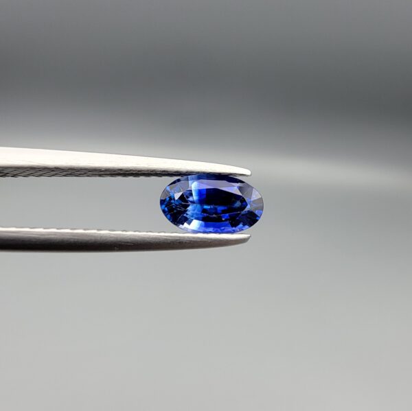 ​Studio close-up photograph of a 0.91 carat natural oval-shaped blue sapphire loose gemstone resting on a textured white surface, showcasing blue facets and a shiny finish.