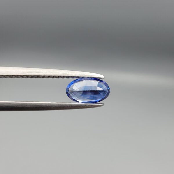 ​Studio close-up photograph of a 0.91 carat natural oval-shaped blue sapphire loose gemstone resting on a textured white surface, showcasing blue facets and a shiny finish.