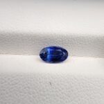 ​Studio close-up photograph of a 0.91 carat natural oval-shaped blue sapphire loose gemstone resting on a textured white surface, showcasing blue facets and a shiny finish.