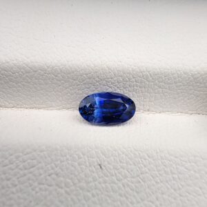 ​Studio close-up photograph of a 0.91 carat natural oval-shaped blue sapphire loose gemstone resting on a textured white surface, showcasing blue facets and a shiny finish.
