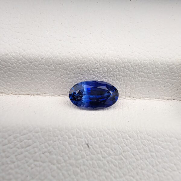 ​Studio close-up photograph of a 0.91 carat natural oval-shaped blue sapphire loose gemstone resting on a textured white surface, showcasing blue facets and a shiny finish.
