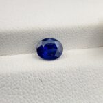 Studio photograph held in steel tweezers showcasing a 1.14 carat natural oval-shaped blue sapphire loose gemstone with deep blue facets and a high-shine finish.