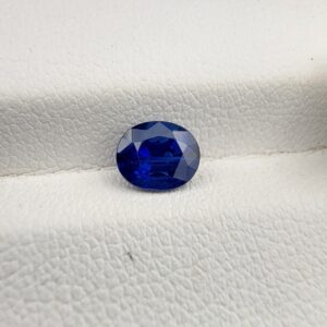 Studio photograph held in steel tweezers showcasing a 1.14 carat natural oval-shaped blue sapphire loose gemstone with deep blue facets and a high-shine finish.