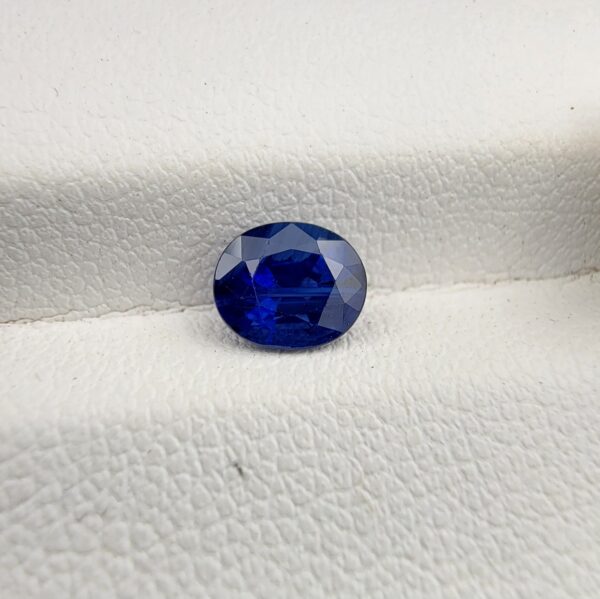 Studio photograph held in steel tweezers showcasing a 1.14 carat natural oval-shaped blue sapphire loose gemstone with deep blue facets and a high-shine finish.