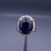 Studio close-up photograph of a handcrafted men's 925 sterling silver ring featuring a massive 19.72 carat oval blue sapphire center stone surrounded by a zircon halo and detailed with seven carved wheel motifs along the shank.