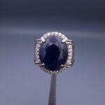 Studio close-up photograph of a handcrafted men's 925 sterling silver ring featuring a massive 19.72 carat oval blue sapphire center stone surrounded by a zircon halo and detailed with seven carved wheel motifs along the shank.