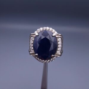 Studio close-up photograph of a handcrafted men's 925 sterling silver ring featuring a massive 19.72 carat oval blue sapphire center stone surrounded by a zircon halo and detailed with seven carved wheel motifs along the shank.