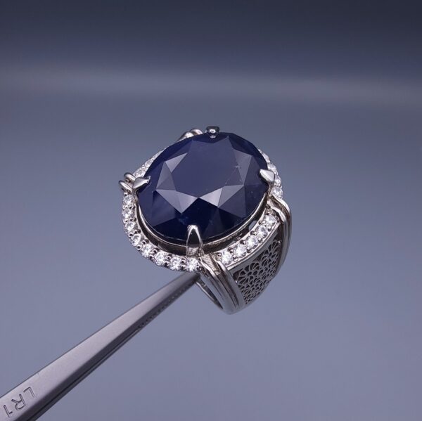 Studio close-up photograph of a handcrafted men's 925 sterling silver ring featuring a massive 19.72 carat oval blue sapphire center stone surrounded by a zircon halo and detailed with seven carved wheel motifs along the shank.