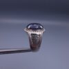 Studio close-up photograph of a handcrafted men's 925 sterling silver ring featuring a massive 19.72 carat oval blue sapphire center stone surrounded by a zircon halo and detailed with seven carved wheel motifs along the shank.