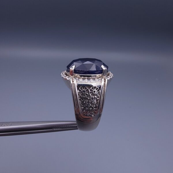 Studio close-up photograph of a handcrafted men's 925 sterling silver ring featuring a massive 19.72 carat oval blue sapphire center stone surrounded by a zircon halo and detailed with seven carved wheel motifs along the shank.