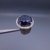Studio close-up photograph of a handcrafted men's 925 sterling silver ring featuring a massive 19.72 carat oval blue sapphire center stone surrounded by a zircon halo and detailed with seven carved wheel motifs along the shank.