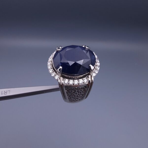 Studio close-up photograph of a handcrafted men's 925 sterling silver ring featuring a massive 19.72 carat oval blue sapphire center stone surrounded by a zircon halo and detailed with seven carved wheel motifs along the shank.