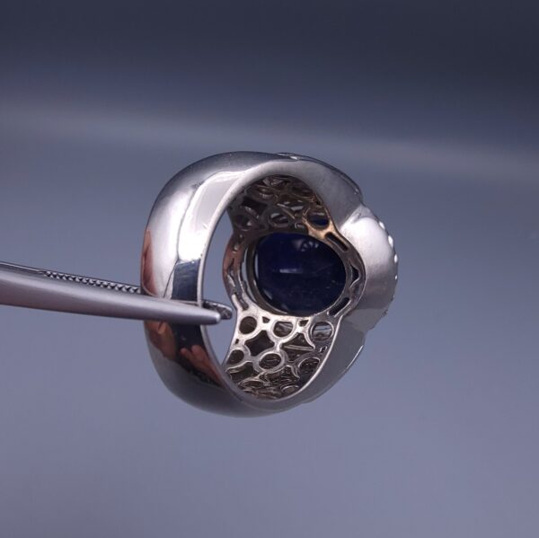 Studio close-up photograph of a handcrafted men's 925 sterling silver ring featuring a massive 19.72 carat oval blue sapphire center stone surrounded by a zircon halo and detailed with seven carved wheel motifs along the shank.