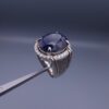 Studio close-up photograph of a handcrafted men's 925 sterling silver ring featuring a massive 19.72 carat oval blue sapphire center stone surrounded by a zircon halo and detailed with seven carved wheel motifs along the shank.