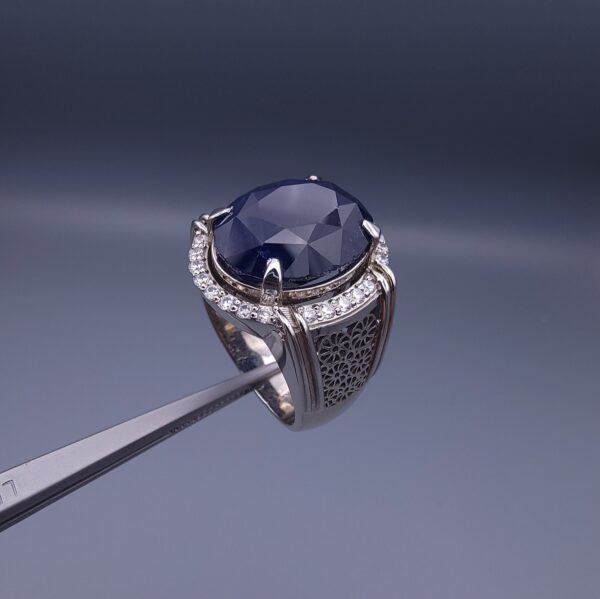 Studio close-up photograph of a handcrafted men's 925 sterling silver ring featuring a massive 19.72 carat oval blue sapphire center stone surrounded by a zircon halo and detailed with seven carved wheel motifs along the shank.