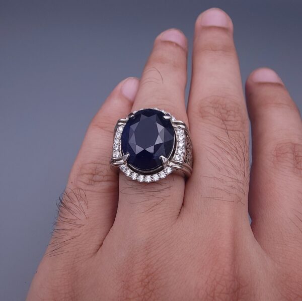 Studio close-up photograph of a handcrafted men's 925 sterling silver ring featuring a massive 19.72 carat oval blue sapphire center stone surrounded by a zircon halo and detailed with seven carved wheel motifs along the shank.
