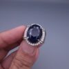Studio close-up photograph of a handcrafted men's 925 sterling silver ring featuring a massive 19.72 carat oval blue sapphire center stone surrounded by a zircon halo and detailed with seven carved wheel motifs along the shank.