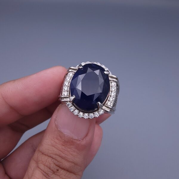 Studio close-up photograph of a handcrafted men's 925 sterling silver ring featuring a massive 19.72 carat oval blue sapphire center stone surrounded by a zircon halo and detailed with seven carved wheel motifs along the shank.