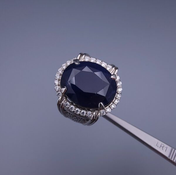 Studio close-up photograph of a handcrafted men's 925 sterling silver ring featuring a massive 19.72 carat oval blue sapphire center stone surrounded by a zircon halo and detailed with seven carved wheel motifs along the shank.