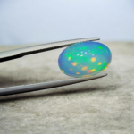 Studio close-up photograph of a 4.16 carat oval cabochon natural Ethiopian Welo opal displaying vibrant green, red, and yellow play-of-color held by tweezers.