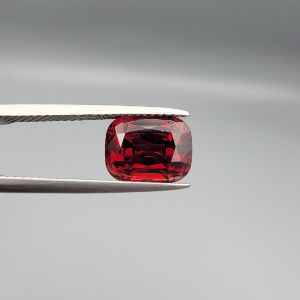 Studio close-up photographs showing the top table face, step-cut pavilion facets, and reverse profile views of a 4.55 carat cushion cut natural almandine garnet loose gemstone held in precision tweezers.