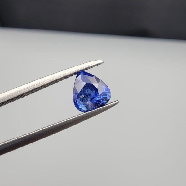 Studio close-up photographs showing the top table and facet profile views of a 1.26 carat pear cut natural blue sapphire loose gemstone held in precision tweezers.