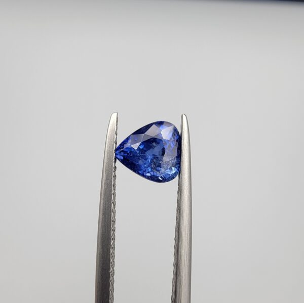 Studio close-up photographs showing the top table and facet profile views of a 1.26 carat pear cut natural blue sapphire loose gemstone held in precision tweezers.
