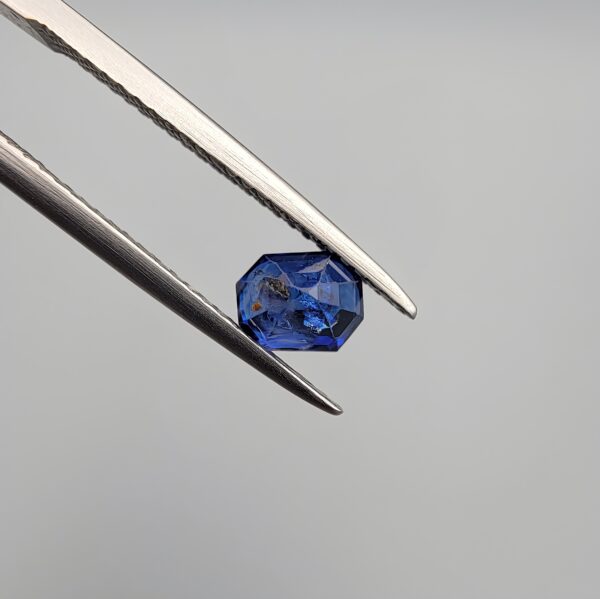 Studio close-up photographs showing the top table and step-cut profile views of a 1.00 carat octagon cut natural blue sapphire loose gemstone held in precision tweezers.