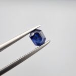 Studio close-up photographs showing the top table and step-cut profile views of a 1.00 carat octagon cut natural blue sapphire loose gemstone held in precision tweezers.