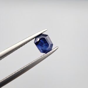 Studio close-up photographs showing the top table and step-cut profile views of a 1.00 carat octagon cut natural blue sapphire loose gemstone held in precision tweezers.