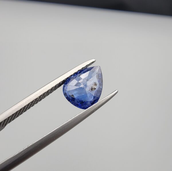 Studio close-up photographs showing the top table view with internal inclusions and reverse facet profile of a 1.41 carat pear cut natural blue sapphire loose gemstone held in precision tweezers.