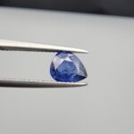 Studio close-up photographs showing the top table view with internal inclusions and reverse facet profile of a 1.41 carat pear cut natural blue sapphire loose gemstone held in precision tweezers.
