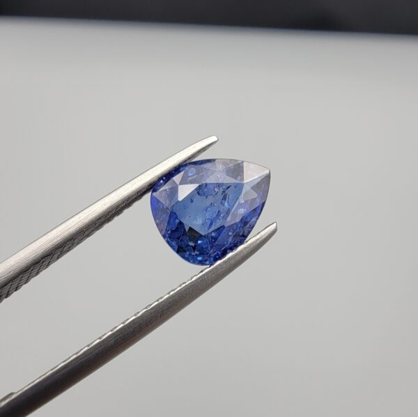 Studio close-up photographs showing the top table view with internal inclusions and reverse facet profile of a 1.41 carat pear cut natural blue sapphire loose gemstone held in precision tweezers.