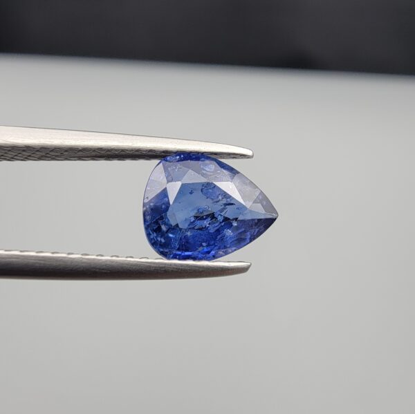 Studio close-up photographs showing the top table view with internal inclusions and reverse facet profile of a 1.41 carat pear cut natural blue sapphire loose gemstone held in precision tweezers.