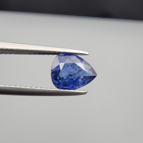Studio close-up photographs showing the top table view with internal inclusions and reverse facet profile of a 1.41 carat pear cut natural blue sapphire loose gemstone held in precision tweezers.