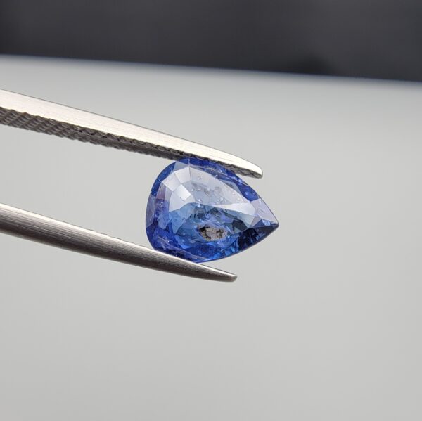 Studio close-up photographs showing the top table view with internal inclusions and reverse facet profile of a 1.41 carat pear cut natural blue sapphire loose gemstone held in precision tweezers.