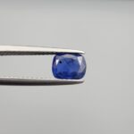 Studio close-up photographs showing the top table face with natural blackish internal inclusions and reverse facet profile of a 1.17 carat cushion cut natural blue sapphire loose gemstone held in precision tweezers.