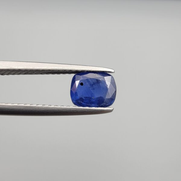 Studio close-up photographs showing the top table face with natural blackish internal inclusions and reverse facet profile of a 1.17 carat cushion cut natural blue sapphire loose gemstone held in precision tweezers.