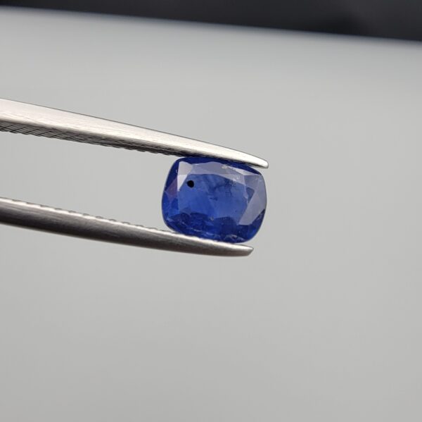 Studio close-up photographs showing the top table face with natural blackish internal inclusions and reverse facet profile of a 1.17 carat cushion cut natural blue sapphire loose gemstone held in precision tweezers.