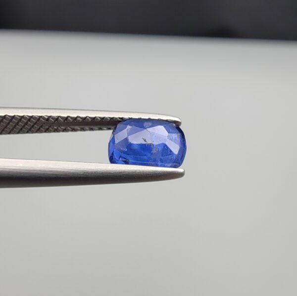 Studio close-up photographs showing the top table face with natural blackish internal inclusions and reverse facet profile of a 1.17 carat cushion cut natural blue sapphire loose gemstone held in precision tweezers.