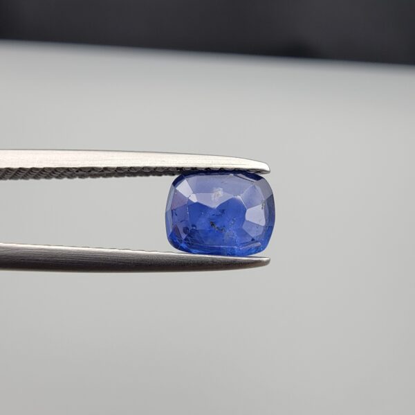 Studio close-up photographs showing the top table face with natural blackish internal inclusions and reverse facet profile of a 1.17 carat cushion cut natural blue sapphire loose gemstone held in precision tweezers.
